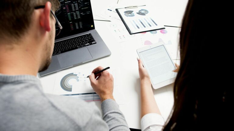 Two business professionals analyzing financial reports using a laptop and tablet in a modern office setting.