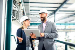 Two people wearing helmets, looking at a screen, in a factory setting.