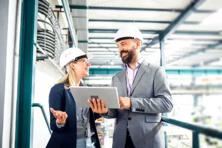 Two people wearing helmets, looking at a screen, in a factory setting.